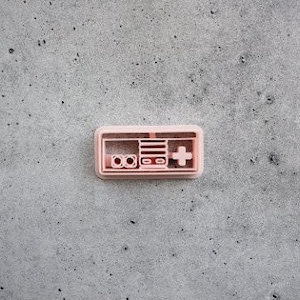 Peut inclure: Un emporte-pièce en plastique rose clair en forme de manette de jeu vidéo rétro. Le design présente un cadre rectangulaire avec deux boutons circulaires à gauche, une zone de console centrale avec des lignes horizontales et une croix directionnelle à droite.