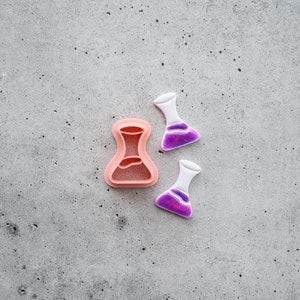 May include: Three pink and white plastic cookie cutters shaped like beakers. The beakers are a light pink color with a purple liquid inside.