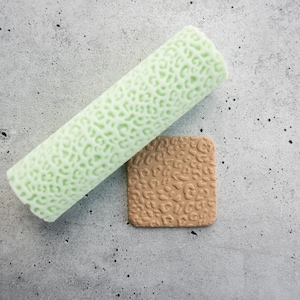 May include: A cylindrical light green rolling pin with a raised leopard print texture and a square tan clay tile featuring the same embossed leopard pattern. The tools are designed for creating consistent textures on soft materials like clay or dough.