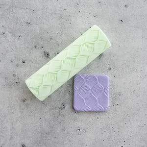 Pode incluir: Um rolo cilíndrico verde-claro com um padrão de textura de folhas repetitivas e um pequeno azulejo quadrado roxo que exibe o mesmo design de folha em relevo, ambos usados para criar impressões em materiais de modelagem macios.