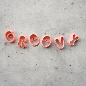 May include: Pink plastic cookie cutters spelling the word "GROOVY" are arranged on a gray speckled surface. The letters are in a rounded, playful font, perfect for baking themed treats. The cutters are a light, pastel pink.