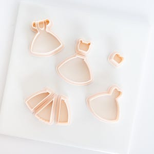 May include: A collection of light peach-colored dress-shaped cookie cutters. The cutters are made of plastic and are in various sizes and styles, including a dress with a flared skirt and a heart-shaped cutter.