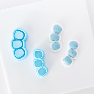 May include: Four blue and white polymer clay earring components and cutters. The components are three stacked rounded shapes with a white border. The cutters are the same shape, in shades of blue. The items are on a white surface.
