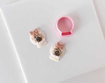 Może przedstawiać: Różowa plastikowa foremka do ciastek w kształcie kuli śnieżnej obok dwóch małych, płaskich, dekoracyjnych kształtów kuli śnieżnej z gliny. Każdy kształt zawiera brązowy domek z piernika, beżową kokardkę i białe kropki imitujące śnieg.
