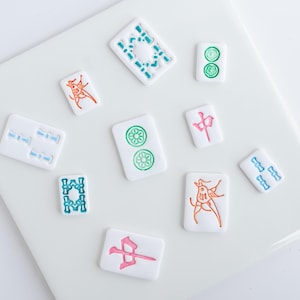 Puede incluir: Un conjunto de fichas de Mahjong blancas con dise&ntilde;os coloridos. Las fichas presentan s&iacute;mbolos en verde, azul, naranja y rosa. Las fichas est&aacute;n dispuestas sobre una superficie blanca, mostrando los componentes del juego.