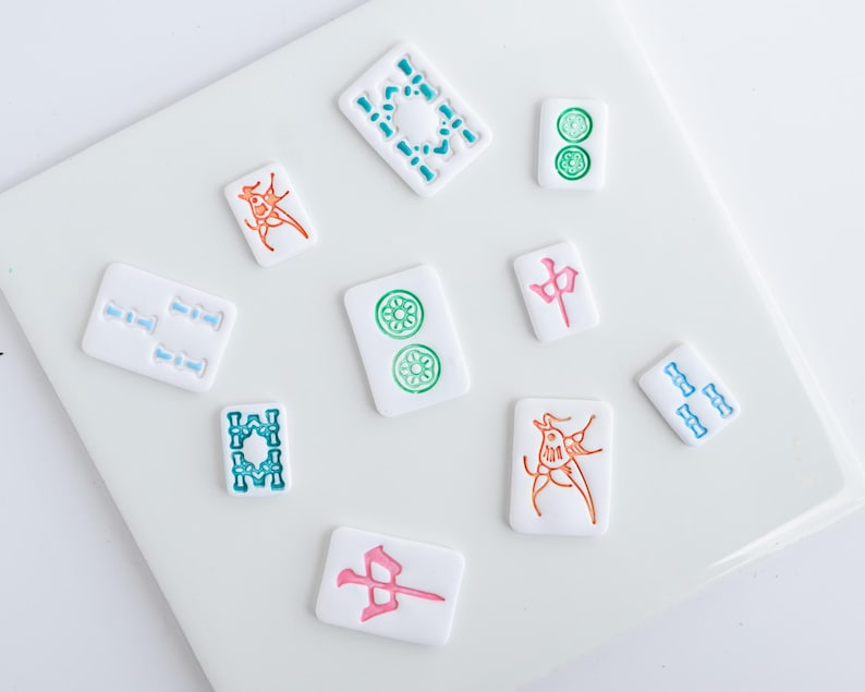 Puede incluir: Un conjunto de fichas de Mahjong blancas con dise&ntilde;os coloridos. Las fichas presentan s&iacute;mbolos en verde, azul, naranja y rosa. Las fichas est&aacute;n dispuestas sobre una superficie blanca, mostrando los componentes del juego.