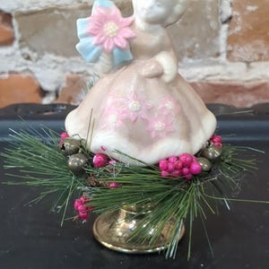 May include: A vintage figurine of a girl in a pink dress holding a pink and blue flower bouquet. The figurine sits atop a brass base surrounded by green pine needles, pink berries, and small silver bells. A charming holiday decoration.
