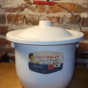 May include: A white enamel chamber pot with a lid and a metal handle featuring a red plastic grip. A label on the front displays the text COLUMBIAN Serving the Home Since 1871, Made in U.S.A. by COLUMBIAN TERRE HAUTE, INDIANA, and a graphic illustration.