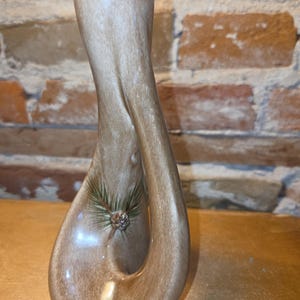 May include: A tall, twisted ceramic vase with a glossy, light brown wood-grain finish. The vase features a central opening and is decorated with a small, painted graphic of a pine cone and green pine needles on the lower front section.