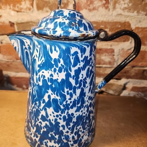 May include: A blue and white speckled enamel coffee pot featuring a matching lid with a small handle, a curved spout, and a black metal handle attached to the side. The surface displays a marbled pattern with visible signs of wear and rust.