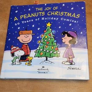 May include: A Peanuts Christmas book cover with a blue background and white stars. The cover features Charlie Brown, Snoopy, Lucy, and a decorated Christmas tree. Text includes "The Joy of A Peanuts Christmas" and "50 Years of Holiday Comics!"