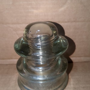 May include: A clear glass electrical insulator with a rounded top and a tiered, bell-shaped design. The glass has a slight green tint and a glossy finish. The insulator is designed to prevent electrical current from flowing through the support structure.