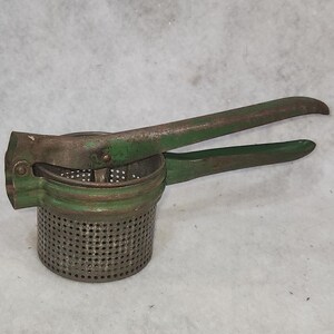 May include: An antique potato ricer with a perforated metal basket and green painted handles. The metal shows signs of age and wear, with a rustic, vintage aesthetic. The handles are designed for easy pressing.