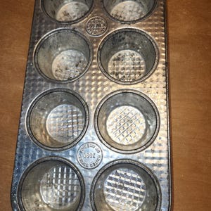 May include: Vintage metal muffin tin with eight circular molds. The tin is a dull silver color with signs of wear and use. The base of each mold has a textured pattern. The brand name "UVENEX" is visible.