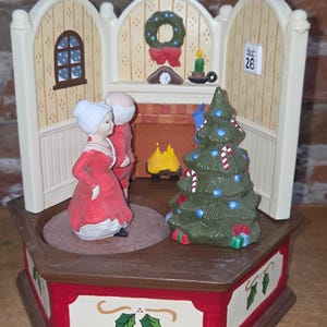 May include: A Christmas-themed musical figurine featuring an elderly couple in red clothing standing before a fireplace. The scene includes a decorated green Christmas tree with candy canes, a wall calendar showing December 26, and a wreath.