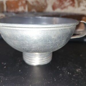 May include: A vintage metal funnel with a wide bowl and a threaded spout. It has a small handle and is made of a silver-colored metal. The funnel is likely used for transferring liquids or dry ingredients.