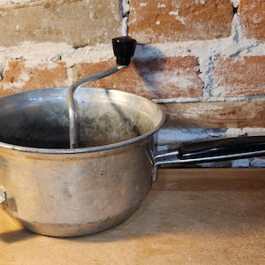 May include: A silver-colored metal food mill featuring a round basin, a side handle with a black plastic grip, and a top-mounted rotating crank with a black knob. The device includes a small metal support tab on the side opposite the handle.
