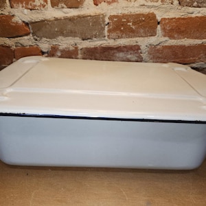 May include: A rectangular, white enamelware baking dish with a matching lid. The dish has a black trim and is set against a brick wall background. The dish is approximately 12 inches long.