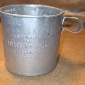May include: A vintage silver-toned metal measuring cup with a handle. The exterior is embossed with the text COMPLIMENTS OF JAVA COFFEE MILLS F.J. PETRU CHICAGO. The interior rim features embossed measurement markings including 3 OZ.