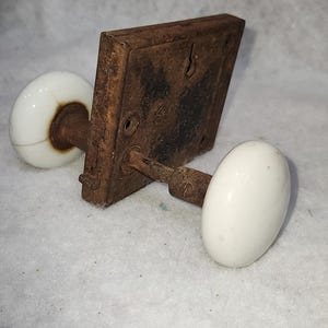 May include: Antique door hardware set featuring a square, heavily rusted metal plate with two white porcelain oval doorknobs. The hardware shows signs of age and wear, with a vintage aesthetic.