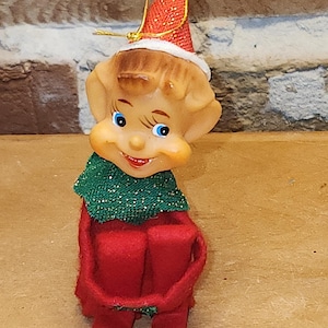 May include: A vintage Christmas elf figurine. The elf has a smiling face with blue eyes, a red and white hat, and a green collar. The elf is wearing a red outfit and has a seated pose. A festive holiday decoration.