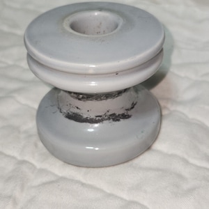 May include: A light gray ceramic insulator with a cylindrical shape. The insulator has a circular top with a central hole, a ridged middle section, and a rounded base. The surface shows some wear and discoloration.