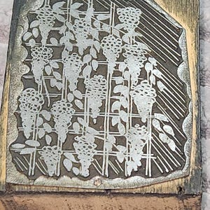 May include: A vintage printing block with a floral design. The metal plate features a detailed pattern of flowers and leaves, set against a grid background. The block is mounted on a wooden base, showing signs of age and use.