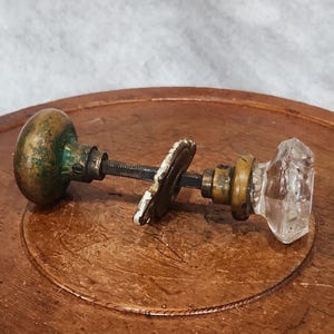 May include: Antique door knobs with a brass and clear glass design. One knob is a rounded brass shape with green patina, the other is a faceted clear glass. The knobs are connected by a metal rod and a decorative plate.