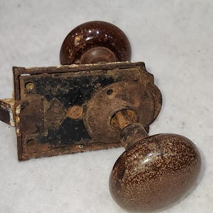 May include: An antique door knob set with a brown ceramic knob and a matching oval-shaped knob. The metal hardware is heavily rusted, showing signs of age and wear. The set includes a rectangular backplate and a spindle.