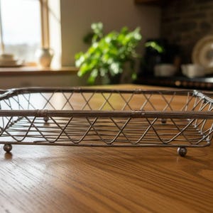 May include: A rectangular silver-toned metal wire basket featuring a cross-hatched side pattern, two integrated handles on the ends, and four small spherical feet. The structure is made of thin, ribbed metal rods arranged in a grid design.