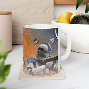 May include: White ceramic mug featuring a detailed illustration of a space battle scene with a soldier in white and blue armor holding a blaster. The background shows spaceships, planets, and an orange and blue sky. The mug is on a beige coaster.