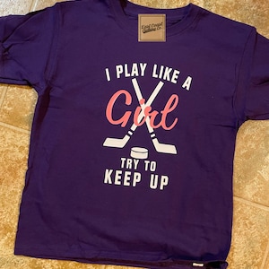 May include: Purple t-shirt with a white graphic of hockey sticks crossed over a puck. The text reads "I play like a Girl try to keep up".