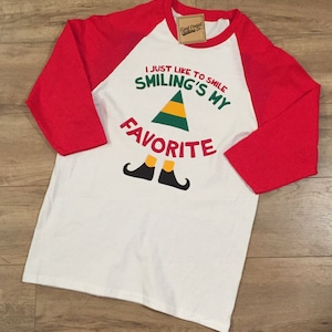 May include: White and red baseball style t-shirt with a green and yellow elf hat and the text "I just like to smile, Smiling's my favourite".