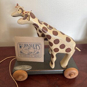 May include: A vintage pull-along toy featuring two giraffes with brown spots and a cream-colored body. The giraffes stand on a green base with wooden wheels. A small cardboard sign reads "WORKSHOPS" and "DR. GERALD E. HENN".