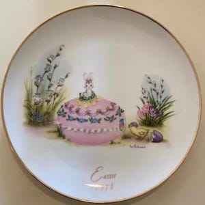 Puede incluir: Plato decorativo de Pascua con borde dorado. El plato presenta un diseño en forma de huevo rosa con un conejito encima, un pollito y la palabra "Easter" con el año 1998.