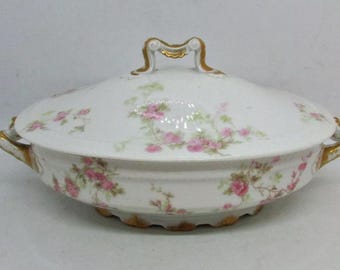 May include: A white porcelain serving dish with a lid, featuring a delicate floral pattern of pink roses and green leaves. The dish has gold accents on the rim and handles.