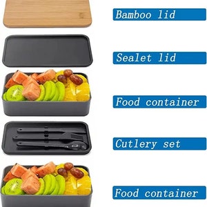 May include: A gray and black lunchbox set with two food containers, a cutlery set, a black sealable lid, and a bamboo lid. The lunchbox is filled with fruit, including watermelon, kiwi, and oranges.