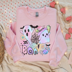May include: A pink sweatshirt with a colorful graphic design featuring ghosts, spiders, bats, and the word "BOO".