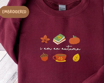 May include: A maroon sweatshirt with embroidered autumn-themed graphics, including a leaf, books, a pumpkin, an acorn, a cup of tea, and a leaf. The text "i am an autumn" is also embroidered on the front.