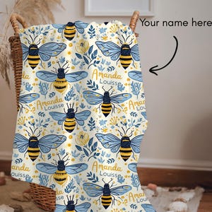 May include: A light-colored blanket with a repeating pattern of blue and yellow bees, flowers, and foliage. The blanket has the text "Amanda Louisse" in a script font. The blanket is folded in a basket.