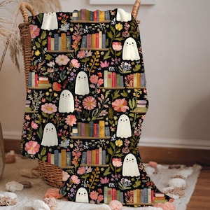 May include: A black throw blanket featuring a repeating pattern of white ghosts, colourful books arranged on shelves, and various pink, yellow, and orange flowers. The design includes multiple horizontal rows of books and scattered floral elements.