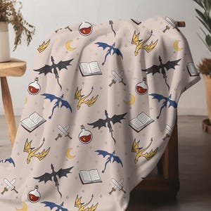 Könnte beinhalten: Eine beige Decke mit einem sich wiederholenden Muster aus schwarzen, blauen und goldenen Drachen, zusammen mit offenen Büchern, Schwertern, Zaubertränken, Monden und Sternen. Die Decke ist über einen Holzstuhl drapiert.