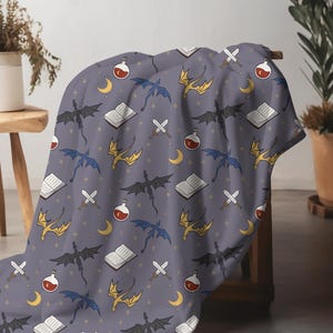 May include: A gray blanket with a repeating pattern of dragons in black, blue, and gold, along with open books, potion bottles, swords, and crescent moons. The blanket is draped over a wooden stool.