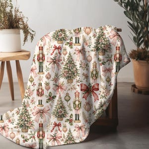 Pode incluir: Uma manta branca com um padrão repetitivo de árvores de Natal em cestos, figuras de quebra-nozes em uniformes vermelhos e verdes, laços cor-de-rosa e ornamentos dourados. O design inclui ramos de azevinho e bagas espalhados pelo tecido.