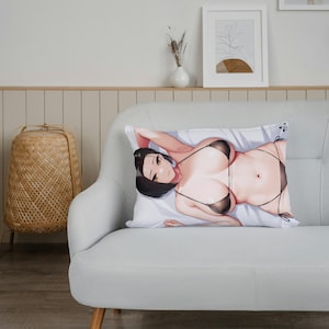 May include: A decorative pillow with a printed image of a woman wearing a black bikini. The pillow is on a light gray couch with wooden legs.
