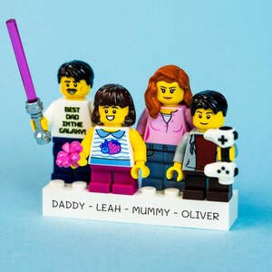 Pode incluir: Uma família de figuras Lego em uma base branca. As figuras incluem um homem segurando um sabre de luz roxo e uma camisa que diz "Best Dad in the Galaxy". A base é rotulada com nomes.