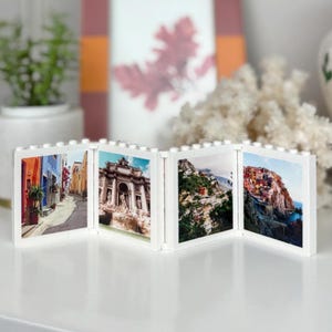 May include: A white photo frame with four separate panels, each displaying a different scenic photograph. The photos are of a European city street, a fountain, a hillside village, and a coastal town.