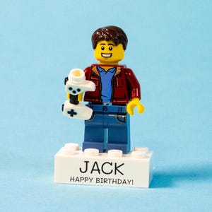 May include: A Lego figurine of a person holding a white video game controller. The figure wears a red jacket, blue shirt, and jeans. The base reads "JACK HAPPY BIRTHDAY!" against a light blue backdrop.