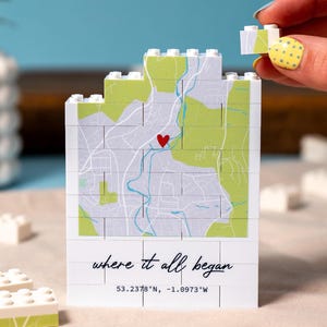 Puede incluir: Un bloque blanco estilo ladrillo con un diseño de mapa, un corazón rojo y el texto "where it all began". El mapa presenta áreas verdes y grises con un río azul. Las coordenadas 53.2378°N, -1.0973°W están impresas debajo. Una mano sostiene un bloque a juego.