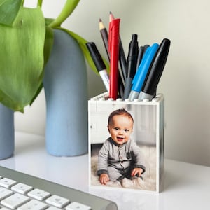 May include: A white block-style pen holder with a photo of a smiling baby. The holder contains various pens and pencils. A keyboard and a blue vase with green leaves are in the background.